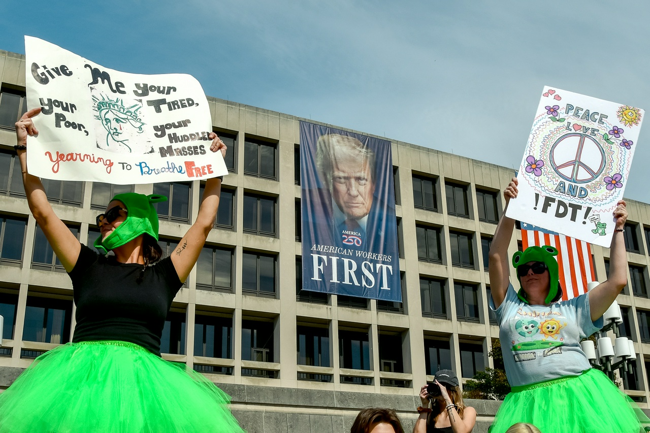 Two women in bright green skirts with protest signs dancing in front of a banner of Donald Trump hanging from the Department of Labor building,