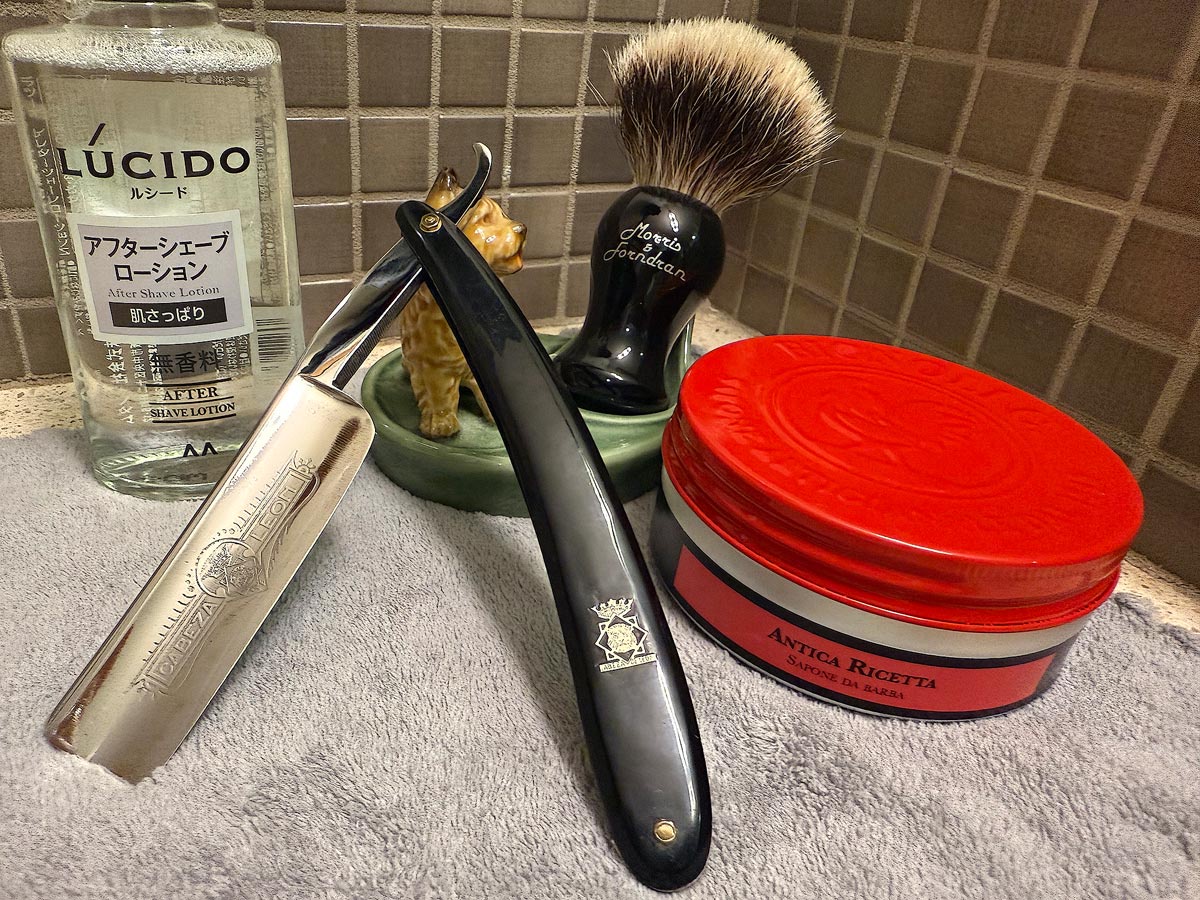 On a gray microfiber towel a straight razor with blade open about 45 degrees leans next to a bottle of aftershave and a jar of shave soap. A shave brush rests bristles-up in a green Wade ceramic pipe rest that features a brown Cairn Terrier.