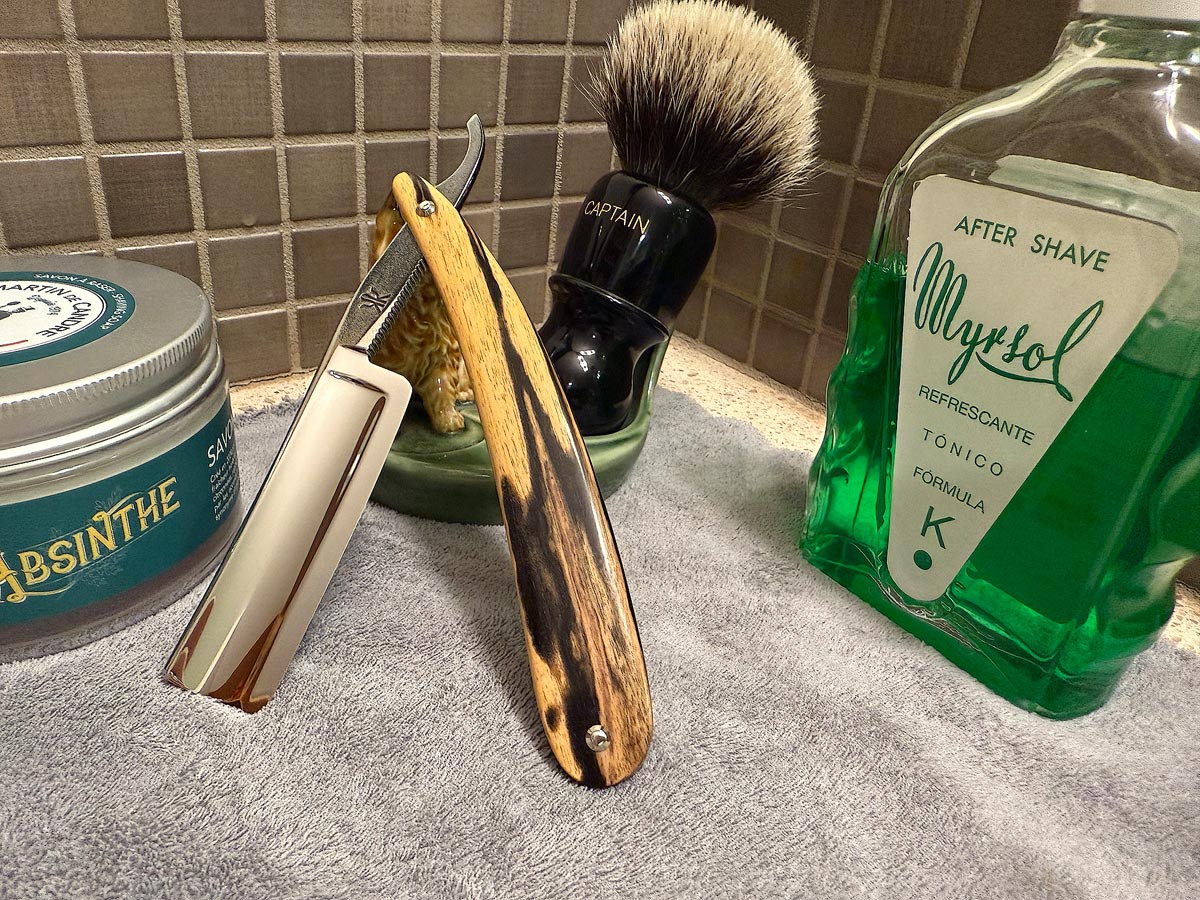 On a gray microfiber towel a straight razor with blade open about 45 degrees leans next to a bottle of aftershave and a jar of shave soap. A shave brush rests bristles-up in a green Wade ceramic pipe rest that features a brown Cairn Terrier.