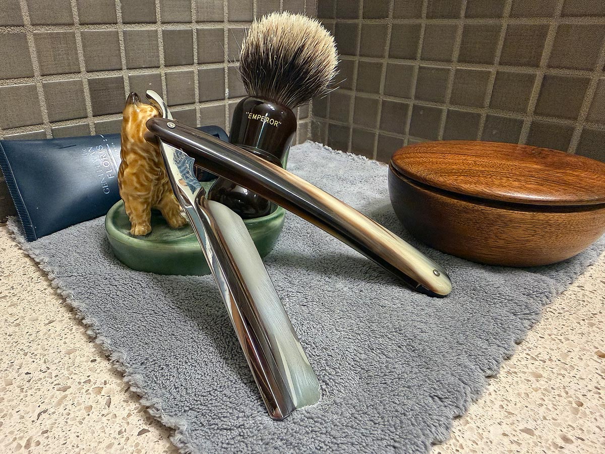 On a gray microfiber towel a straight razor with blade open about 45 degrees leans next to a bottle of aftershave and a jar of shave soap. A shave brush rests bristles-up in a green Wade ceramic pipe rest that features a brown Cairn Terrier.
