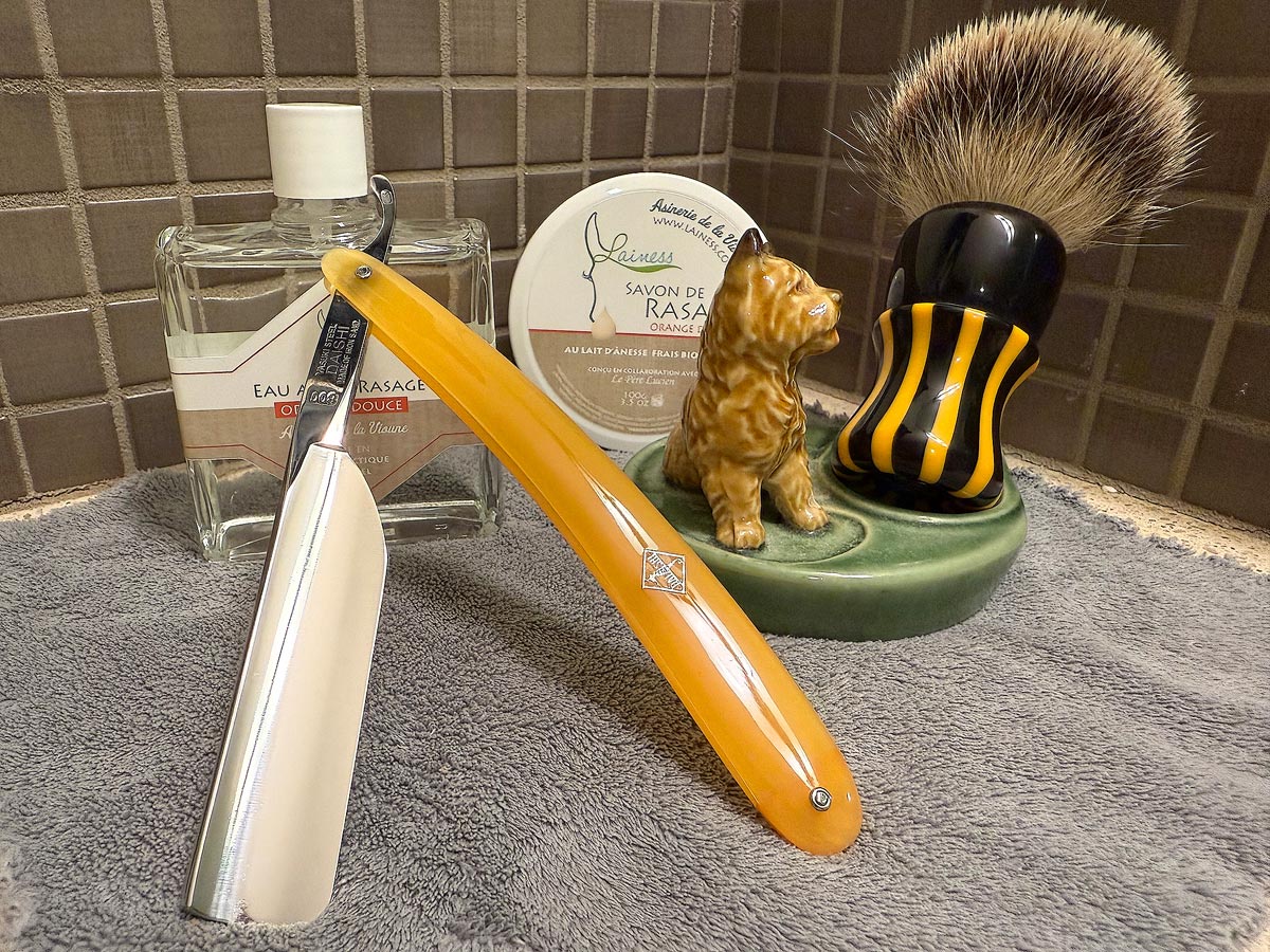 On a gray microfiber towel a straight razor with blade open about 45 degrees leans next to a bottle of aftershave and a jar of shave soap. A shave brush rests bristles-up in a green Wade ceramic pipe rest that features a brown Cairn Terrier.
