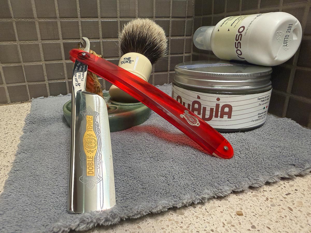 On a gray microfiber towel a straight razor with blade open about 45 degrees leans next to a bottle of aftershave and a jar of shave soap. A shave brush rests bristles-up in a green Wade ceramic pipe rest that features a brown Cairn Terrier.