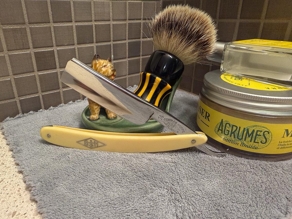 On a gray microfiber towel a straight razor with blade open about 45 degrees leans next to a bottle of aftershave and a jar of shave soap. A shave brush rests bristles-up in a green Wade ceramic pipe rest that features a brown Cairn Terrier.