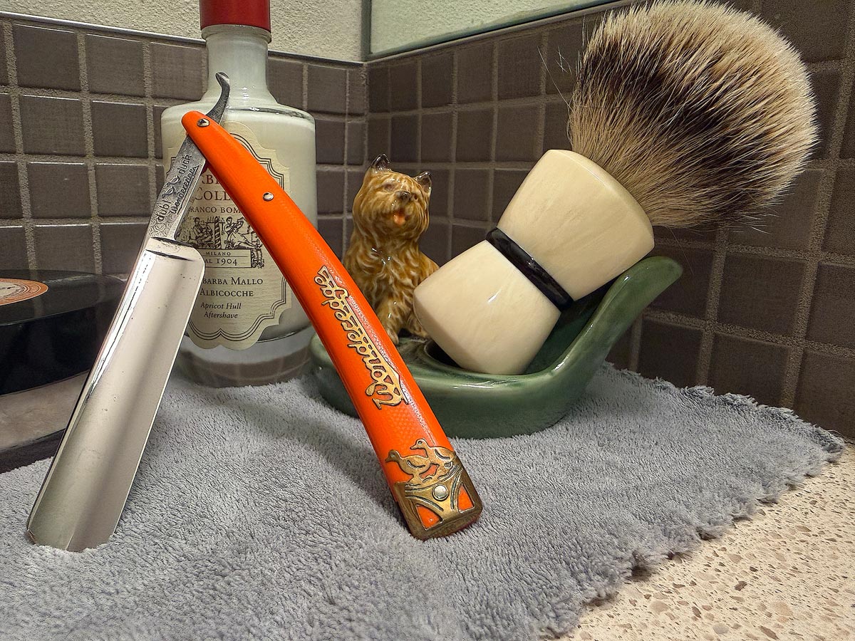 On a gray microfiber towel a straight razor with blade open about 45 degrees leans next to a bottle of aftershave and a jar of shave soap. A shave brush rests bristles-up in a green Wade ceramic pipe rest that features a brown Cairn Terrier.