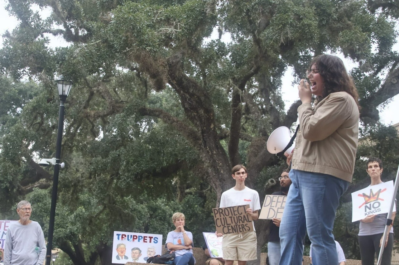 A speaker leads chants on a college campus while protesters hold signs that say, "Protect Cultural Centers" and "No Kings."