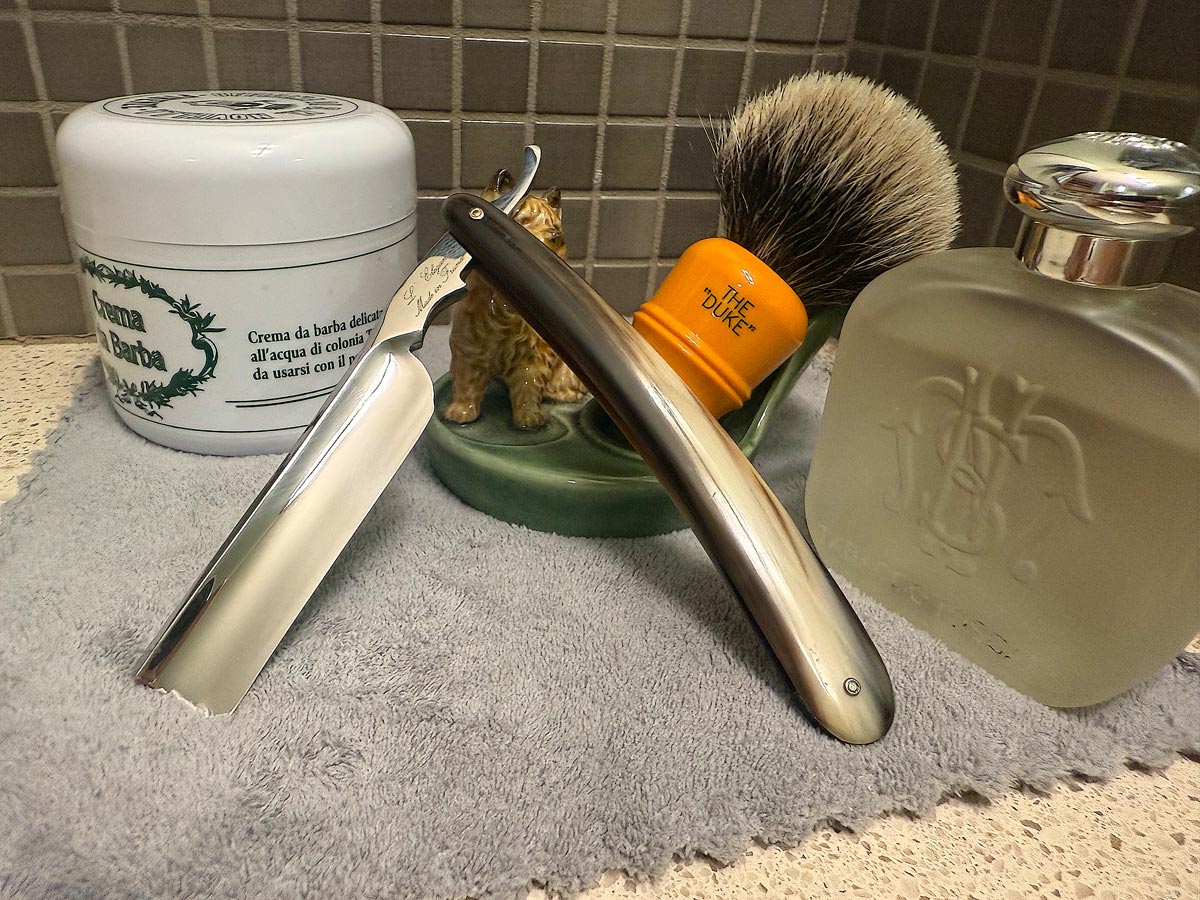 On a gray microfiber towel a straight razor with blade open about 45 degrees leans next to a bottle of aftershave and a jar of shave soap. A shave brush rests bristles-up in a green Wade ceramic pipe rest that features a brown Cairn Terrier.