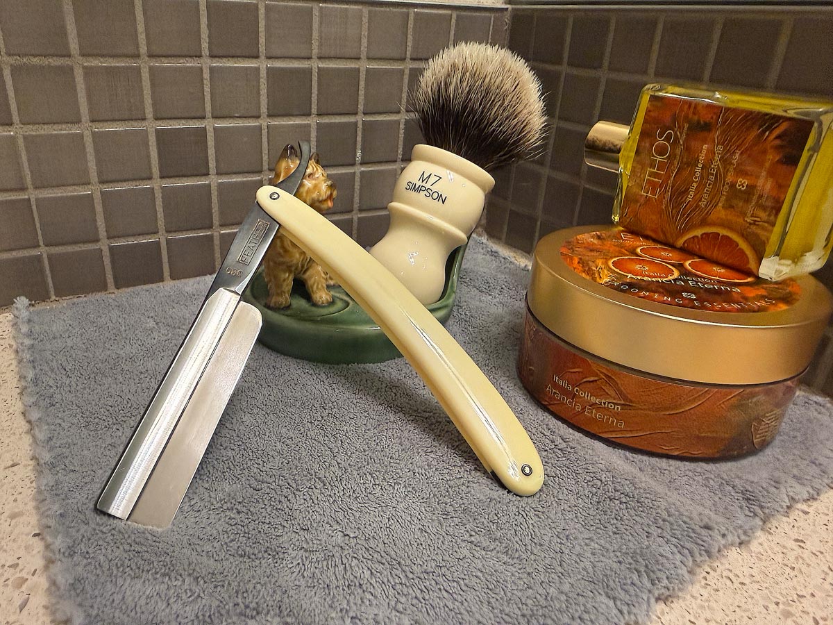 On a gray microfiber towel a straight razor with blade open about 45 degrees leans next to a bottle of aftershave and a jar of shave soap. A shave brush rests bristles-up in a green Wade ceramic pipe rest that features a brown Cairn Terrier.