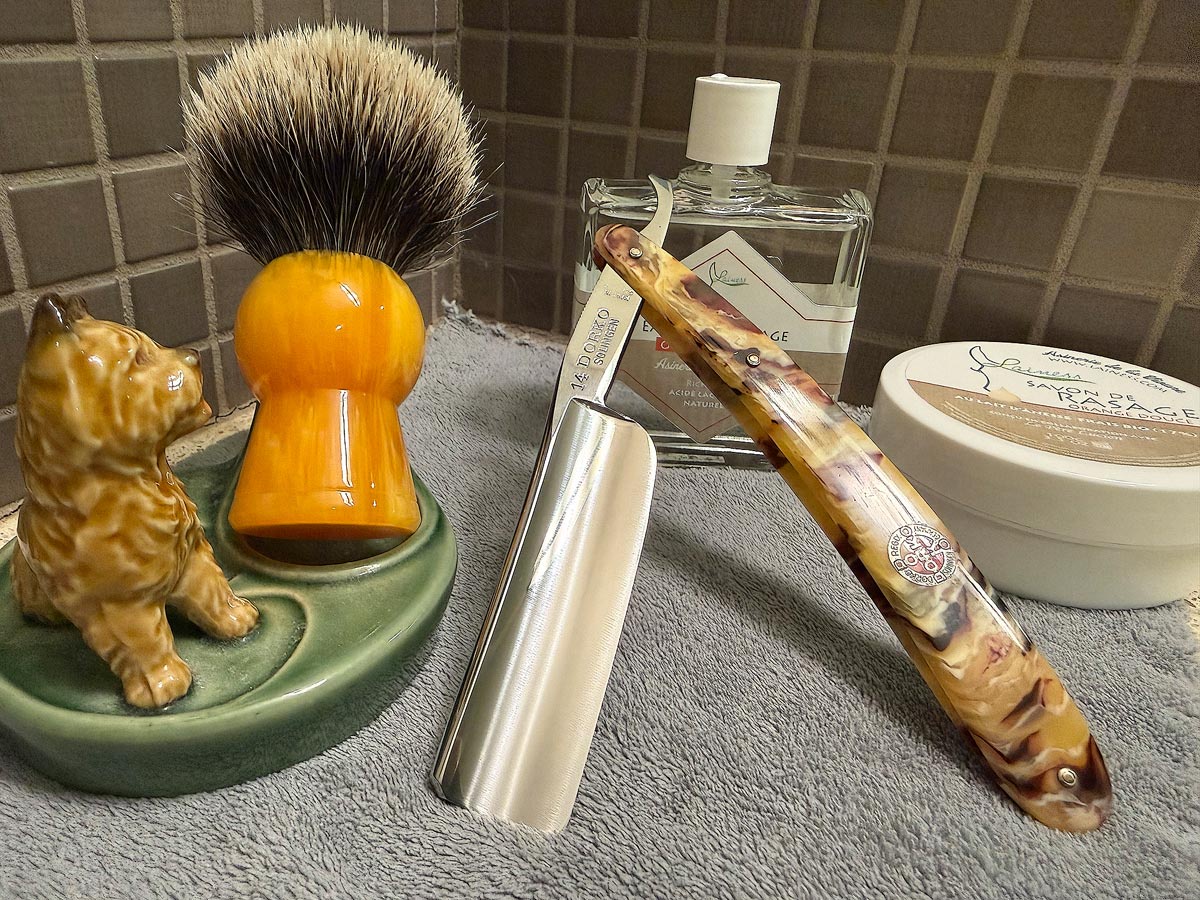 On a gray microfiber towel a straight razor with blade open about 45 degrees leans next to a bottle of aftershave and a jar of shave soap. A shave brush rests bristles-up in a green Wade ceramic pipe rest that features a brown Cairn Terrier.