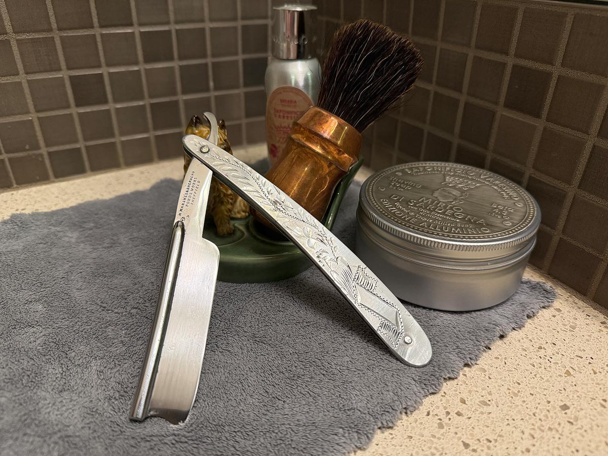 On a gray microfiber towel a straight razor with blade open about 45 degrees leans next to a bottle of aftershave and a jar of shave soap. A shave brush rests bristles-up in a green Wade ceramic pipe rest that features a brown Cairn Terrier.