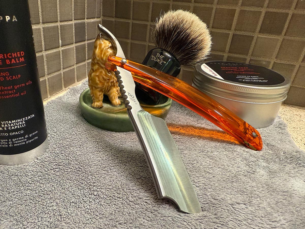 On a gray microfiber towel a straight razor with blade open about 45 degrees leans next to a bottle of aftershave and a jar of shave soap. A shave brush rests bristles-up in a green Wade ceramic pipe rest that features a brown Cairn Terrier.