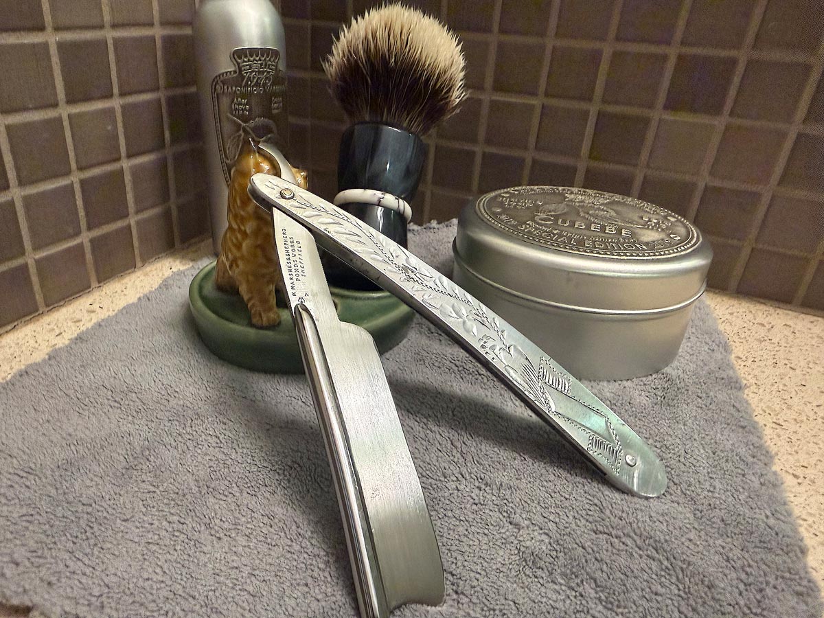On a gray microfiber towel a straight razor with blade open about 45 degrees leans next to a bottle of aftershave and a jar of shave soap. A shave brush rests bristles-up in a green Wade ceramic pipe rest that features a brown Cairn Terrier.