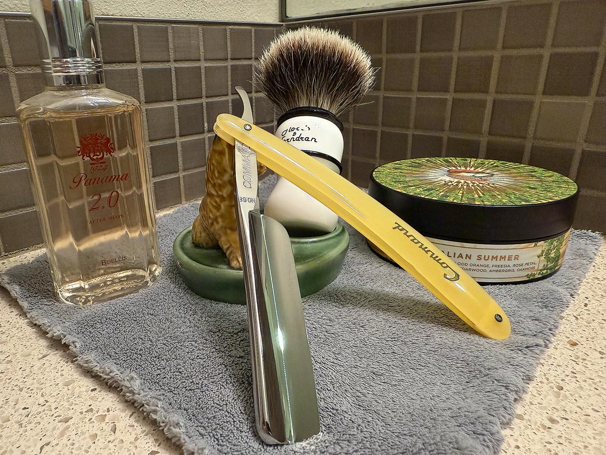 On a gray microfiber towel a straight razor with blade open about 45 degrees leans next to a bottle of aftershave and a jar of shave soap. A shave brush rests bristles-up in a green Wade ceramic pipe rest that features a brown Cairn Terrier.
