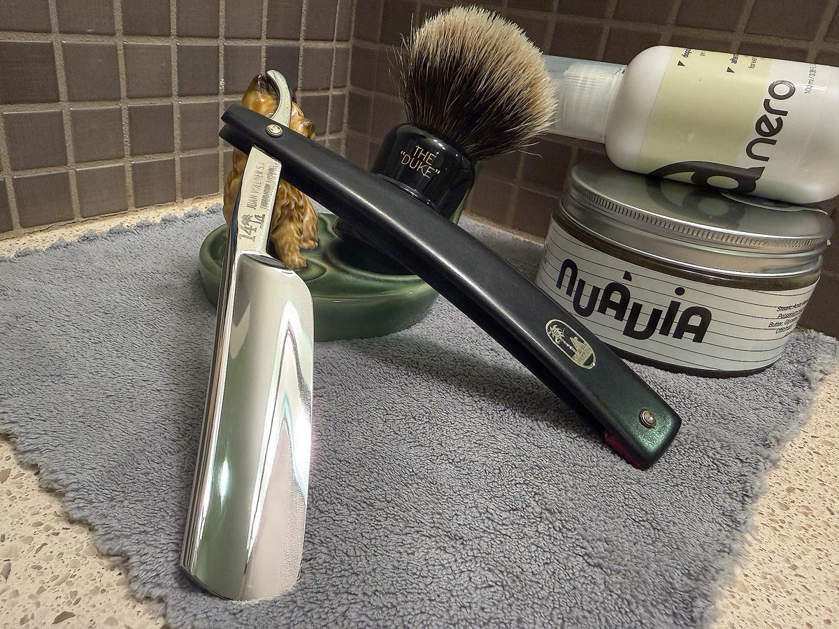 On a gray microfiber towel a straight razor with blade open about 45 degrees leans next to a bottle of aftershave and a jar of shave soap. A shave brush rests bristles-up in a green Wade ceramic pipe rest that features a brown Cairn Terrier.