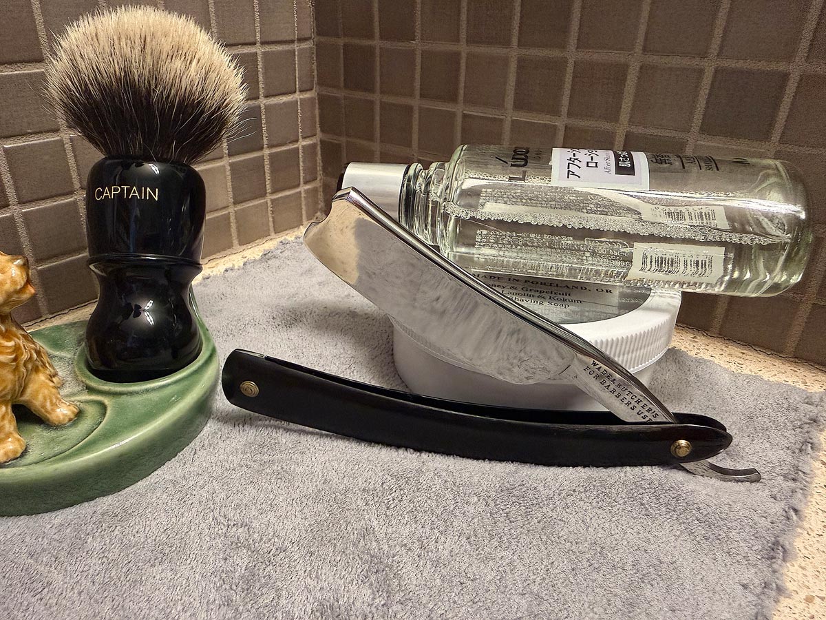 On a gray microfiber towel a straight razor with blade open about 45 degrees leans next to a bottle of aftershave and a jar of shave soap. A shave brush rests bristles-up in a green Wade ceramic pipe rest that features a brown Cairn Terrier.