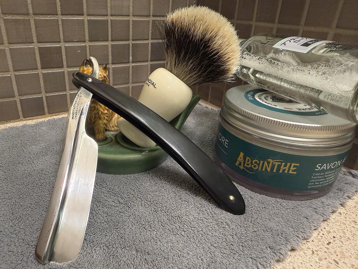 On a gray microfiber towel a straight razor with blade open about 45 degrees leans next to a bottle of aftershave and a jar of shave soap. A shave brush rests bristles-up in a green Wade ceramic pipe rest that features a brown Cairn Terrier.