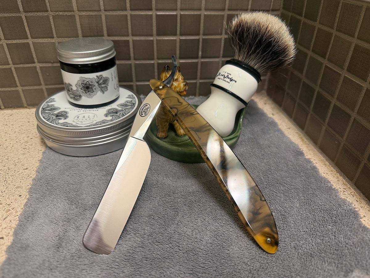 On a gray microfiber towel a straight razor with blade open about 45 degrees leans next to a bottle of aftershave and a jar of shave soap. A shave brush rests bristles-up in a green Wade ceramic pipe rest that features a brown Cairn Terrier.