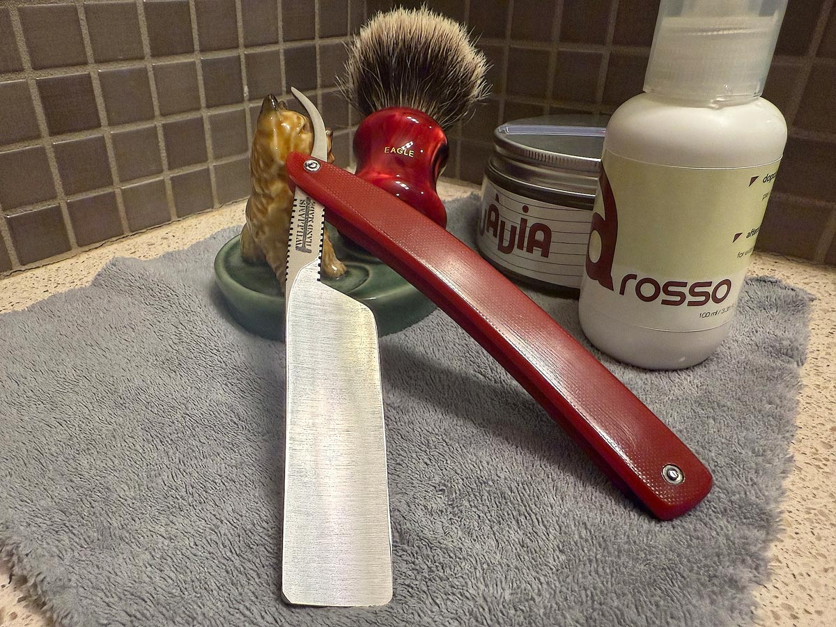 On a gray microfiber towel a straight razor with blade open about 45 degrees leans next to a bottle of aftershave and a jar of shave soap. A shave brush rests bristles-up in a green Wade ceramic pipe rest that features a brown Cairn Terrier.