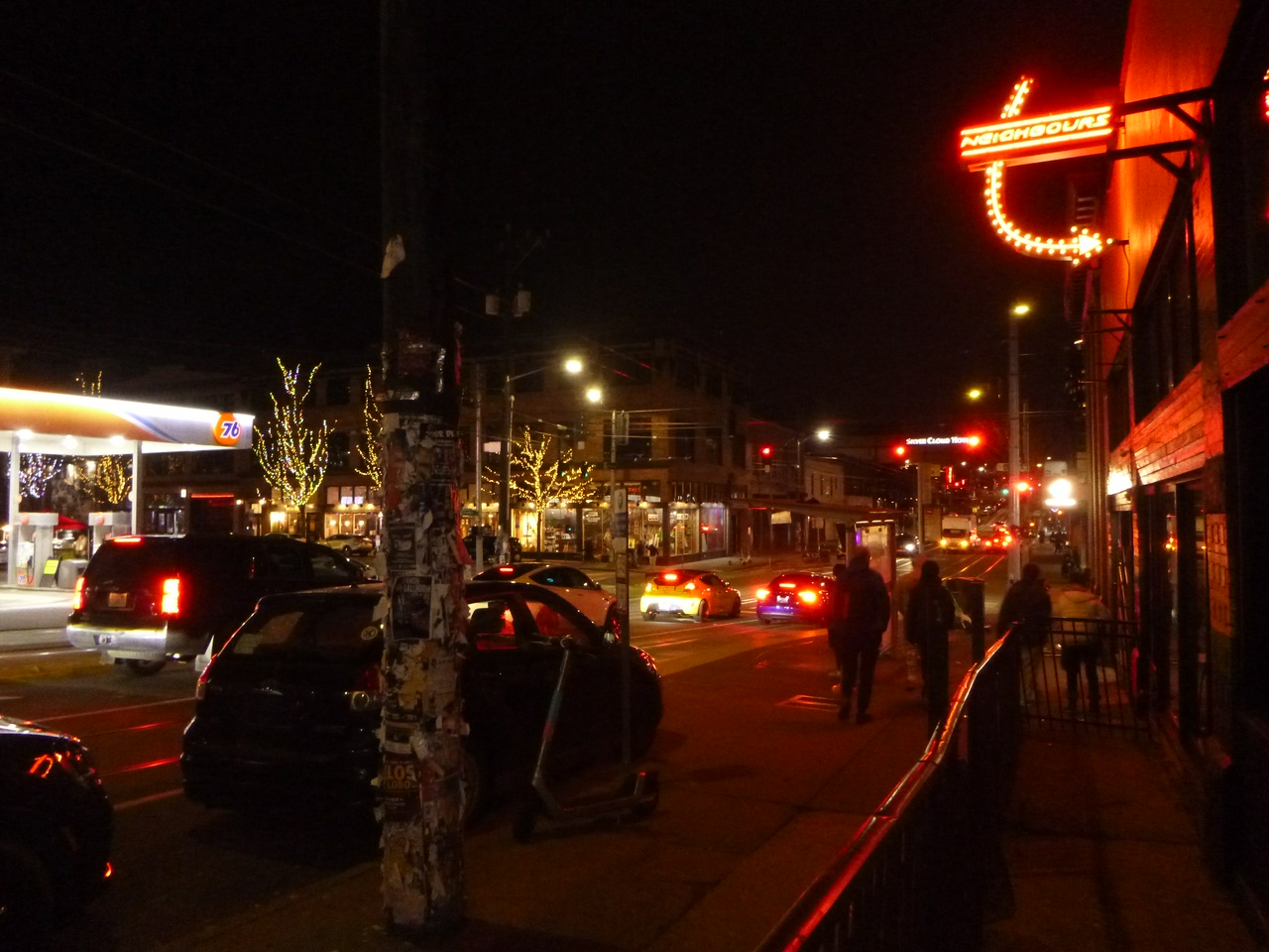 a photo of the intersection of broadway & pike at nighttime, under a pitch-black sky. the street is drenched in a red glow from the car headlights, stop lights, and a bar sign above the sidewalk. a 76 gas station poking out from the left side of the picture sticks out with its bright white lighting.