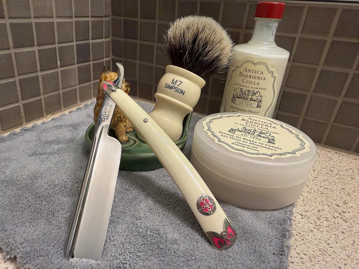 On a gray microfiber towel a straight razor with blade open about 45 degrees leans next to a bottle of aftershave and a jar of shave soap. A shave brush rests bristles-up in a green Wade ceramic pipe rest that features a brown Cairn Terrier.