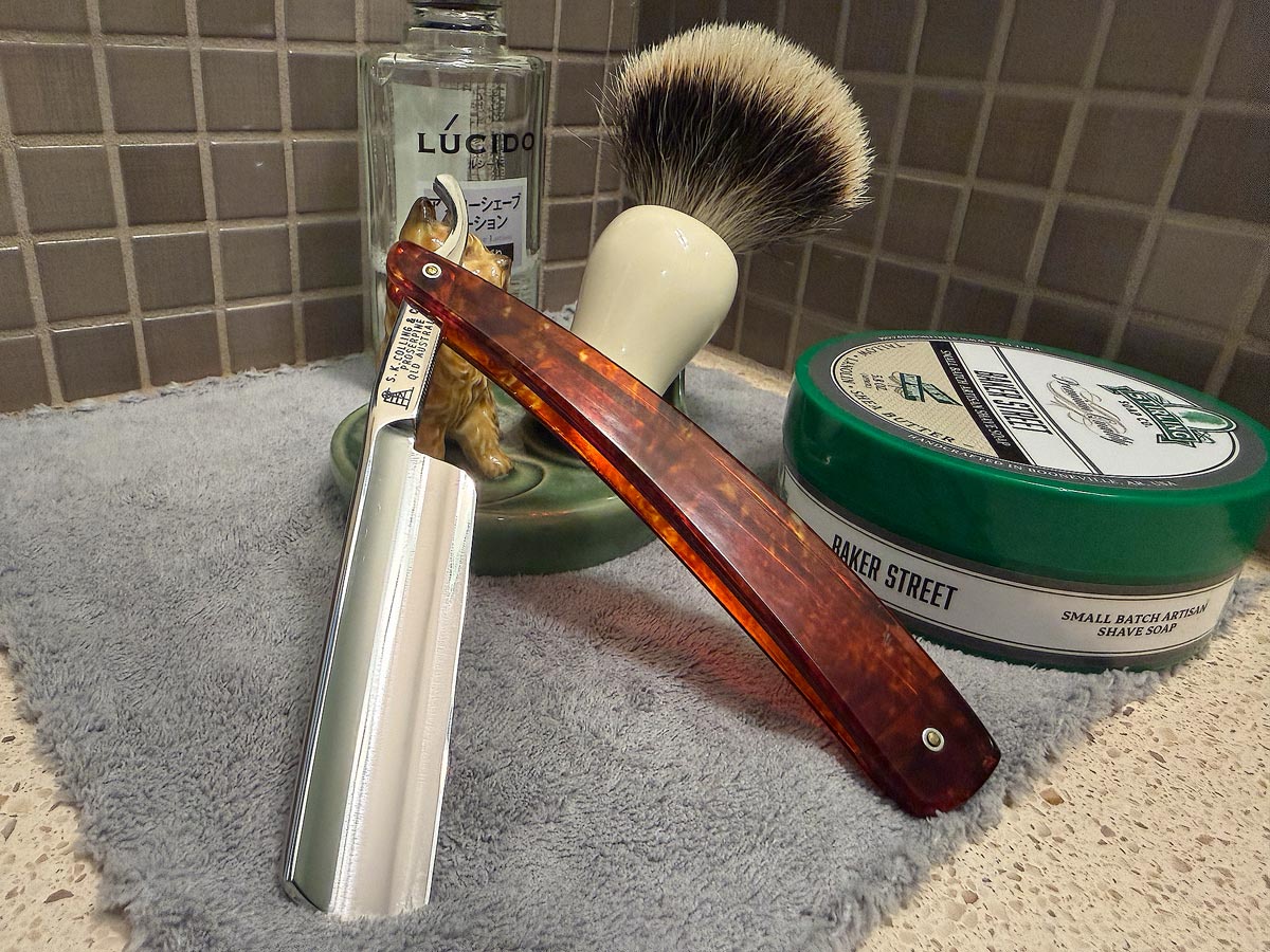 On a gray microfiber towel a straight razor with blade open about 45 degrees leans next to a bottle of aftershave and a jar of shave soap. A shave brush rests bristles-up in a green Wade ceramic pipe rest that features a brown Cairn Terrier.