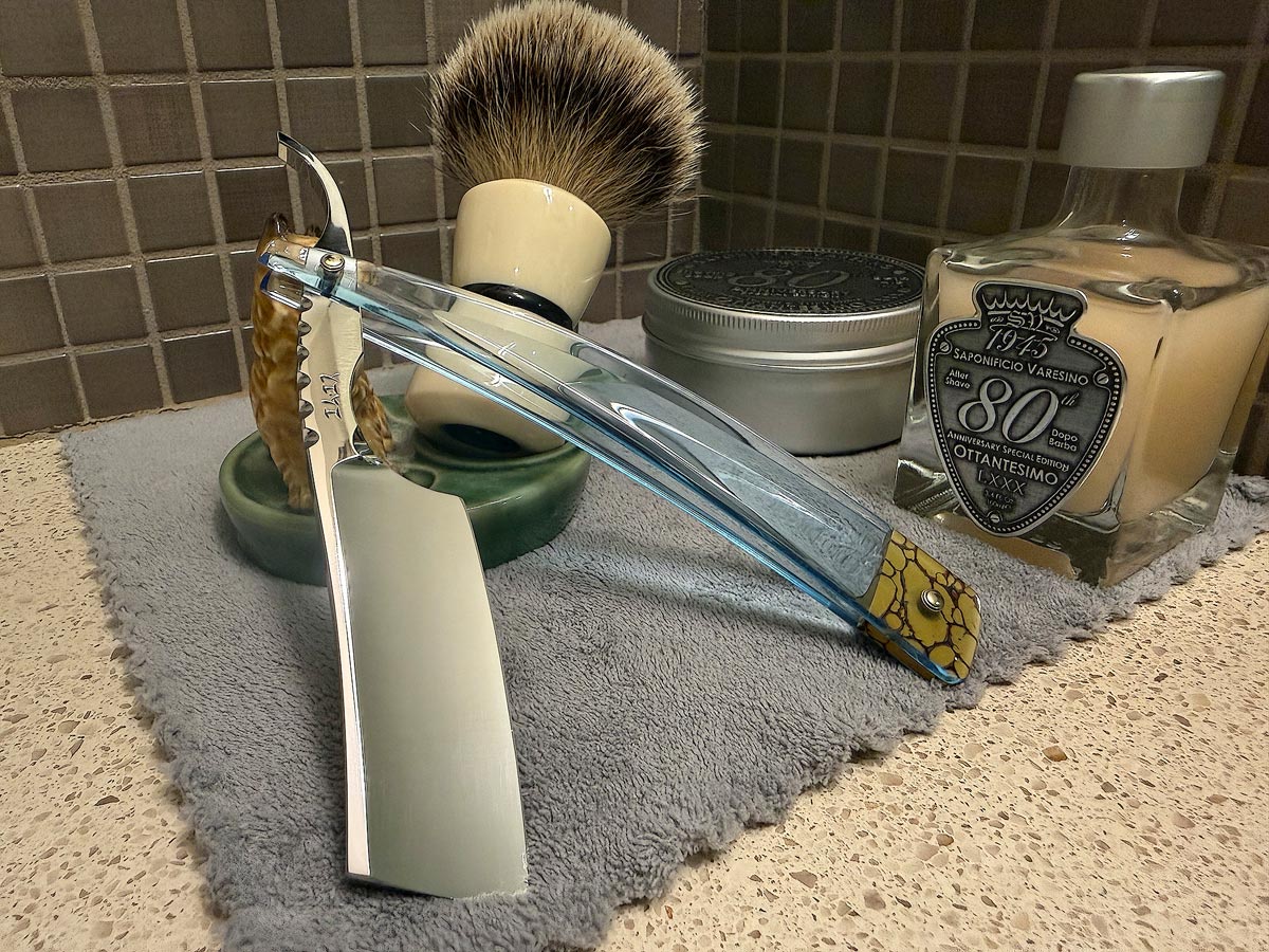 On a gray microfiber towel a straight razor with blade open about 45 degrees leans next to a bottle of aftershave and a jar of shave soap. A shave brush rests bristles-up in a green Wade ceramic pipe rest that features a brown Cairn Terrier.