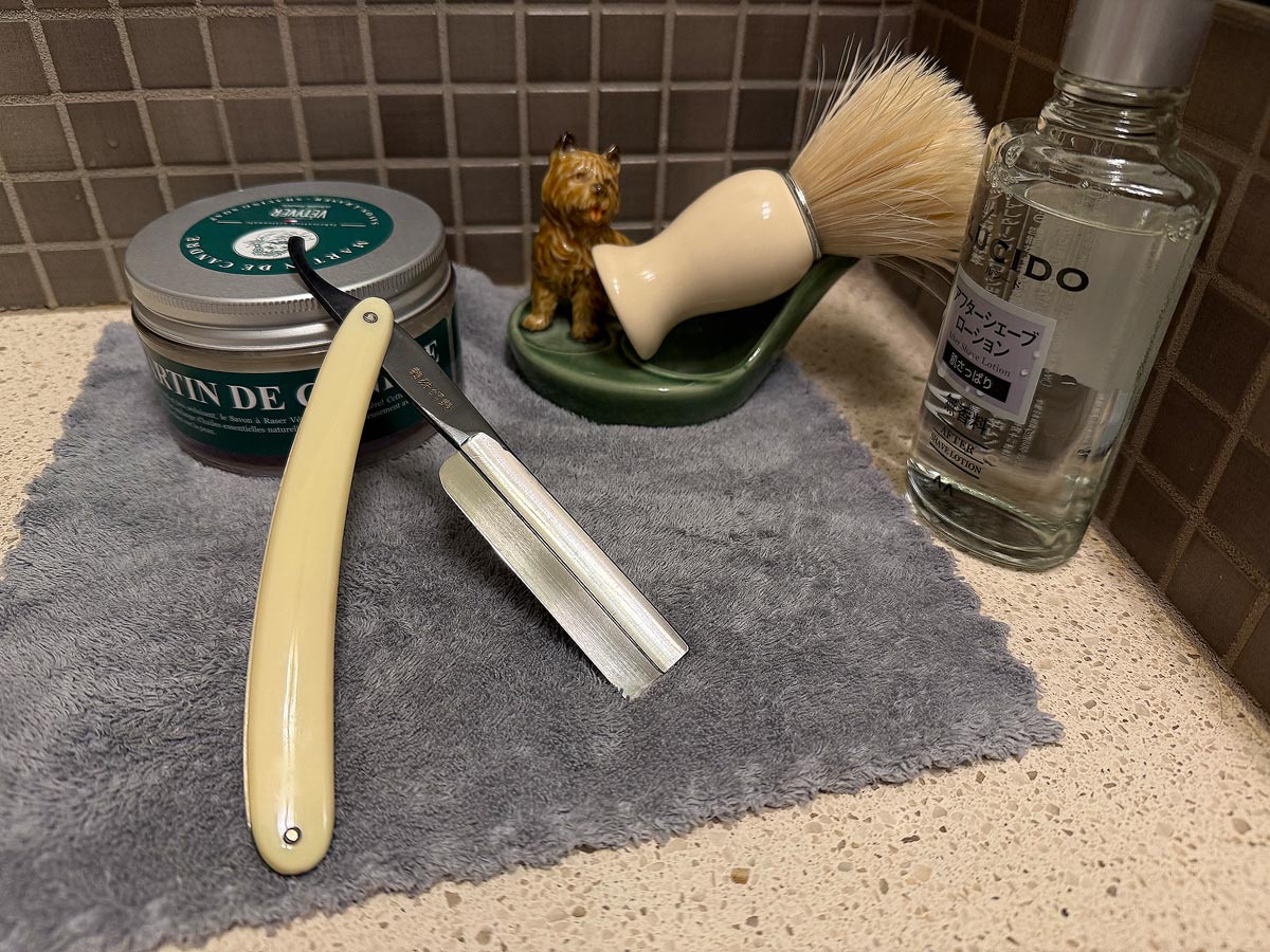 On a gray microfiber towel a straight razor with blade open about 45 degrees leans next to a bottle of aftershave and a jar of shave soap. A shave brush rests bristles-up in a green Wade ceramic pipe rest that features a brown Cairn Terrier.