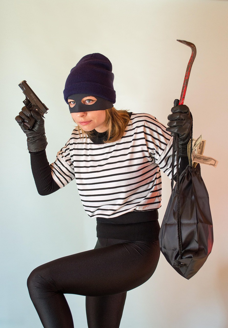 A woman dressed up as a sterotypical thief in a pose that suggest she's sneaking around. She has a black mask that obscures the top of her face. Her hands are in black leather gloves. She holds up a gun with her right hand and a crowbar in her right hand. Her left hand holds up a bag with money sticking out of it. She wears a striped white and black shirt.