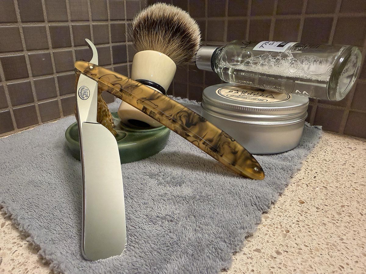 On a gray microfiber towel a straight razor with blade open about 45 degrees leans next to a bottle of aftershave and a jar of shave soap. A shave brush rests bristles-up in a green Wade ceramic pipe rest that features a brown Cairn Terrier.