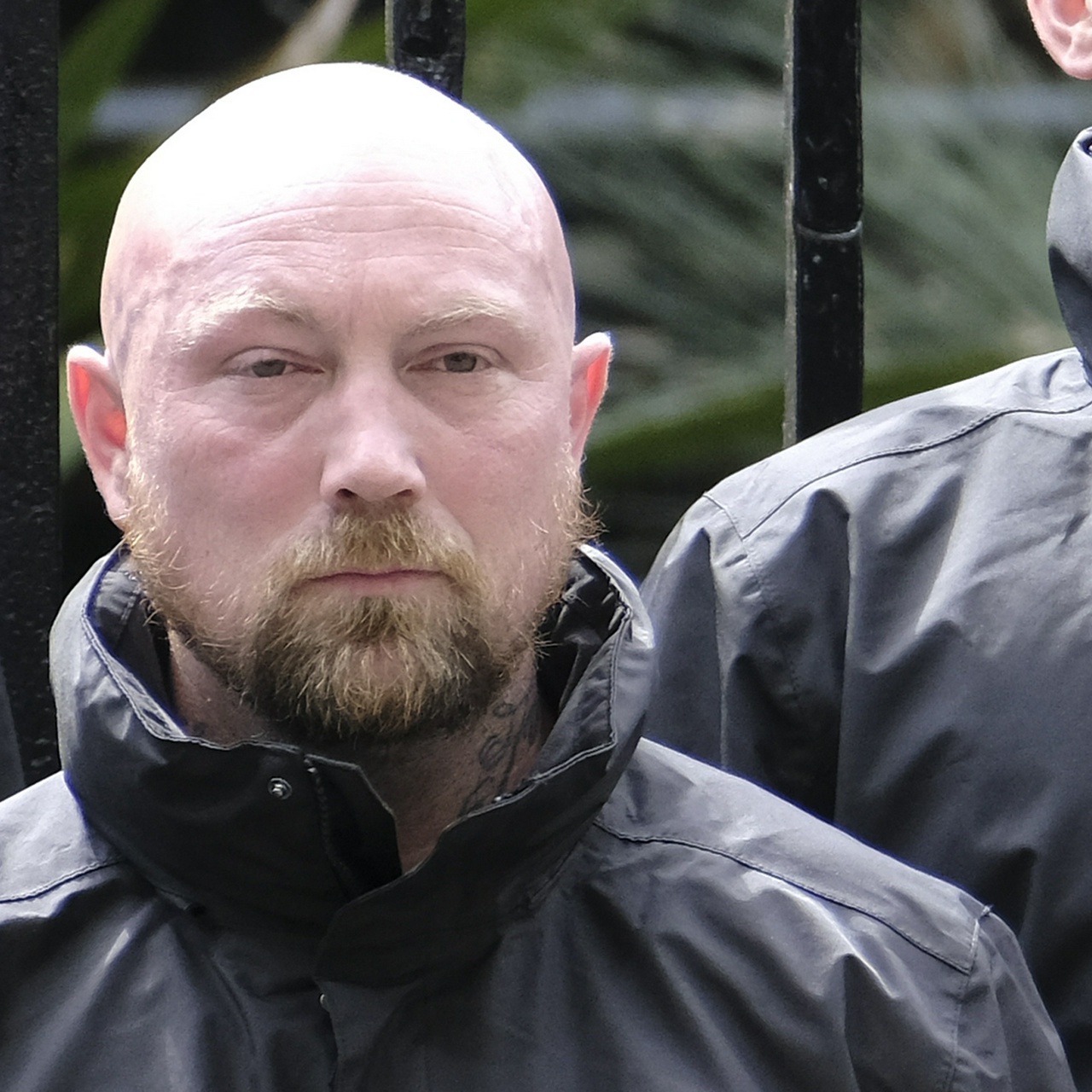 A heavy-lidded bald white man with a very red face and ears, ginger beard and neck tattoos