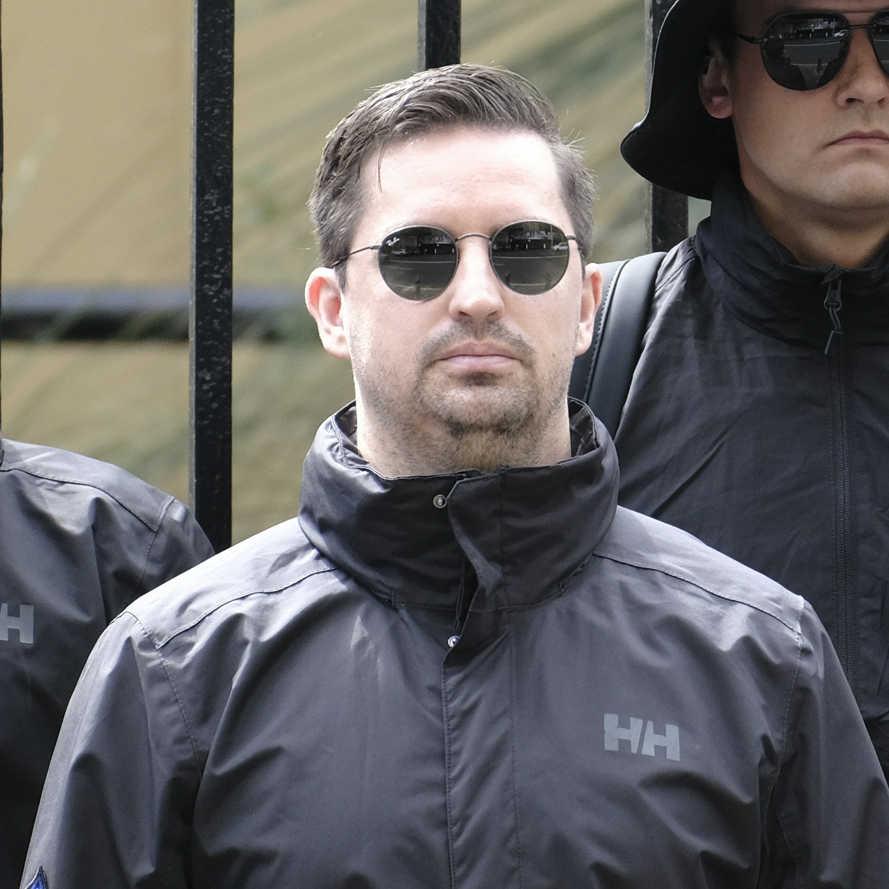 A scruffy, frowning man with a gelled combover and no chin, in round black rayban sunglasses
