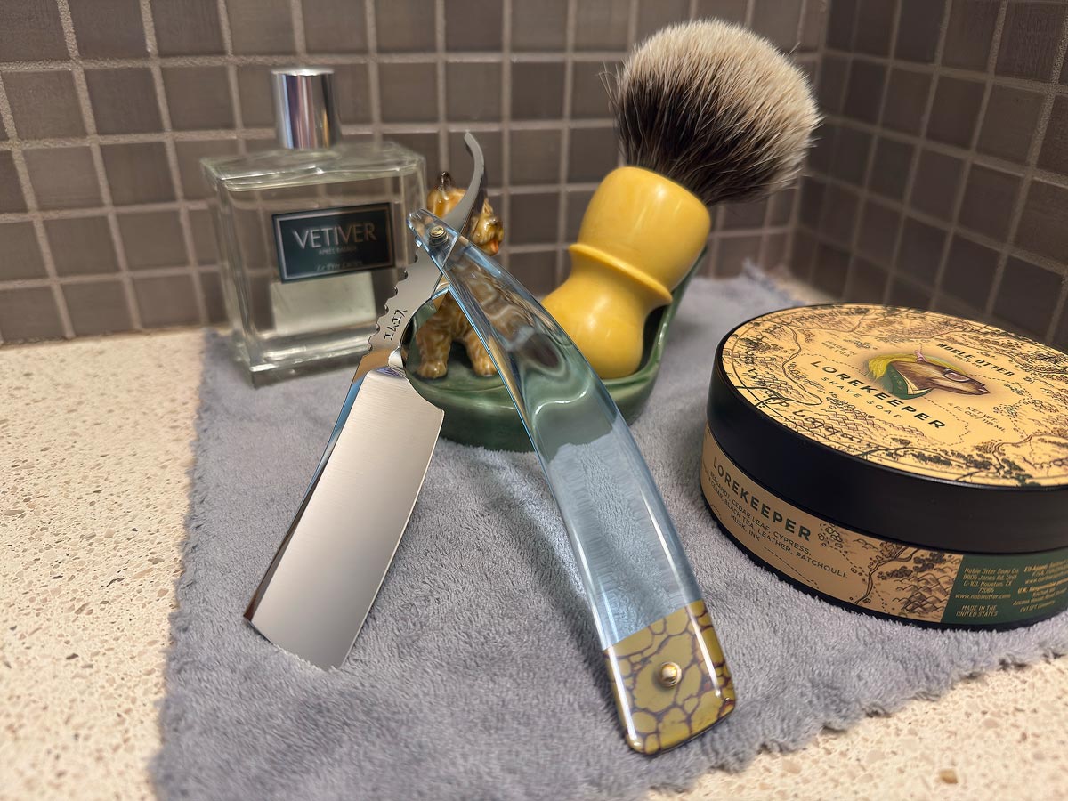 On a gray microfiber towel a straight razor with blade open about 45 degrees leans next to a bottle of aftershave and a jar of shave soap. A shave brush rests bristles-up in a green Wade ceramic pipe rest that features a brown Cairn Terrier.