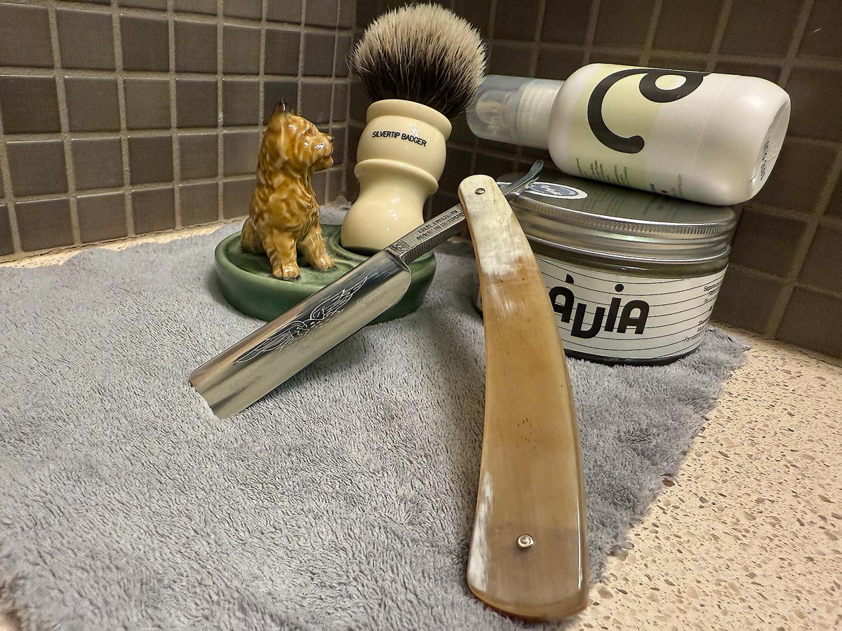 On a gray microfiber towel a straight razor with blade open about 45 degrees leans next to a bottle of aftershave and a jar of shave soap. A shave brush rests bristles-up in a green Wade ceramic pipe rest that features a brown Cairn Terrier.