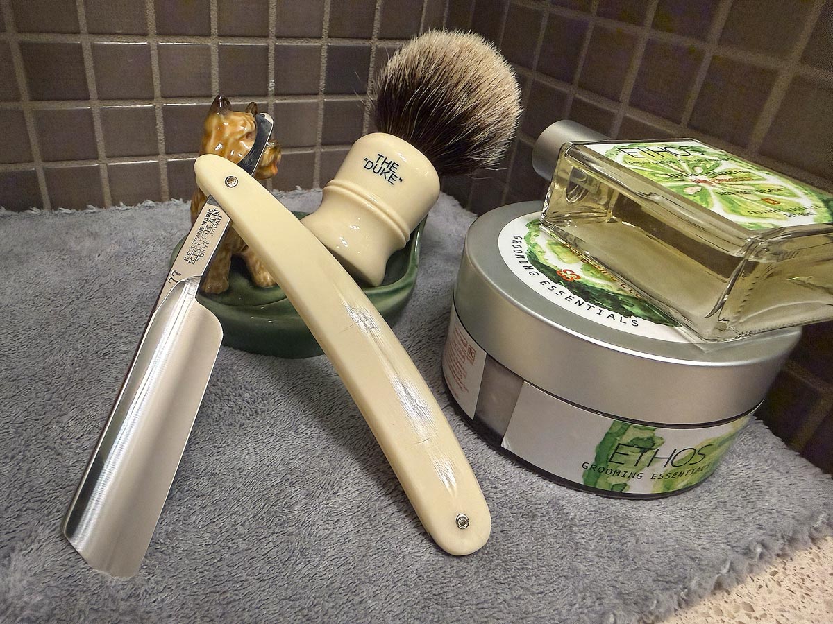 On a gray microfiber towel a straight razor with blade open about 45 degrees leans next to a bottle of aftershave and a jar of shave soap. A shave brush rests bristles-up in a green Wade ceramic pipe rest that features a brown Cairn Terrier.