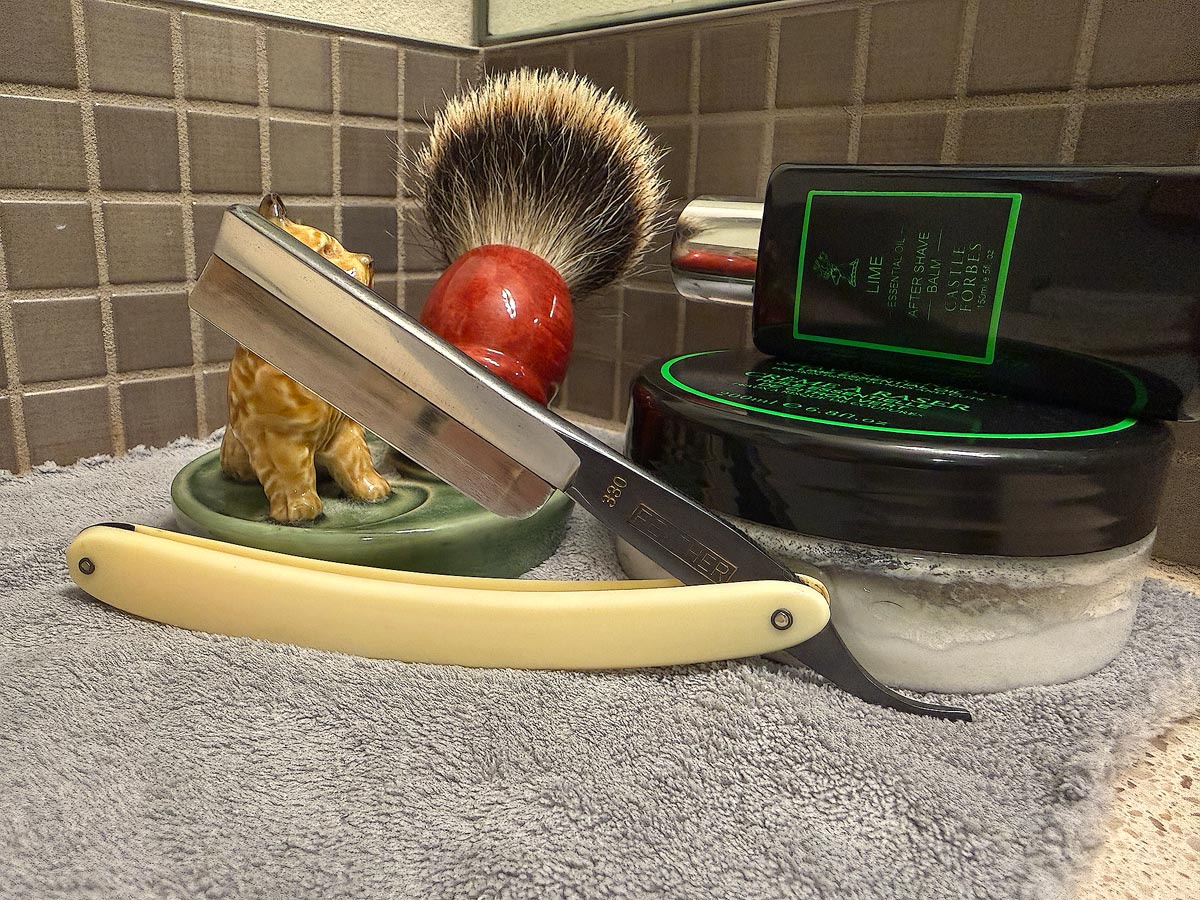 On a gray microfiber towel a straight razor with blade open about 45 degrees leans next to a bottle of aftershave and a jar of shave soap. A shave brush rests bristles-up in a green Wade ceramic pipe rest that features a brown Cairn Terrier.