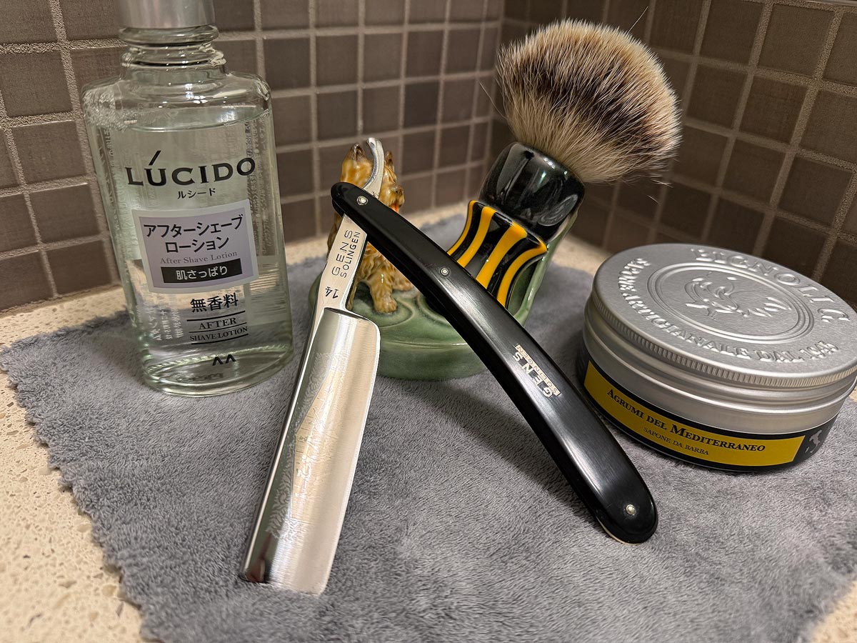 On a gray microfiber towel a straight razor with blade open about 45 degrees leans next to a bottle of aftershave and a jar of shave soap. A shave brush rests bristles-up in a green Wade ceramic pipe rest that features a brown Cairn Terrier.
