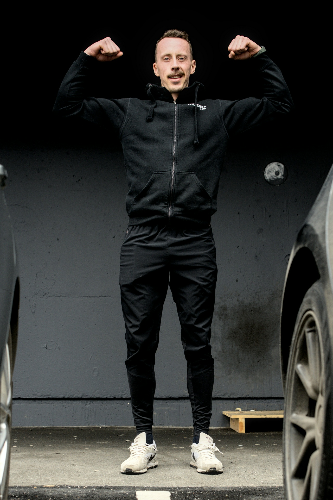 En mann i svart treningsdrakt flexer biceps mot en svart bakgrunn 