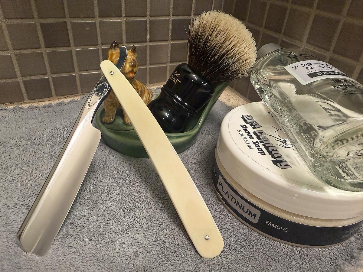 On a gray microfiber towel a straight razor with blade open about 45 degrees leans next to a bottle of aftershave and a jar of shave soap. A shave brush rests bristles-up in a green Wade ceramic pipe rest that features a brown Cairn Terrier.