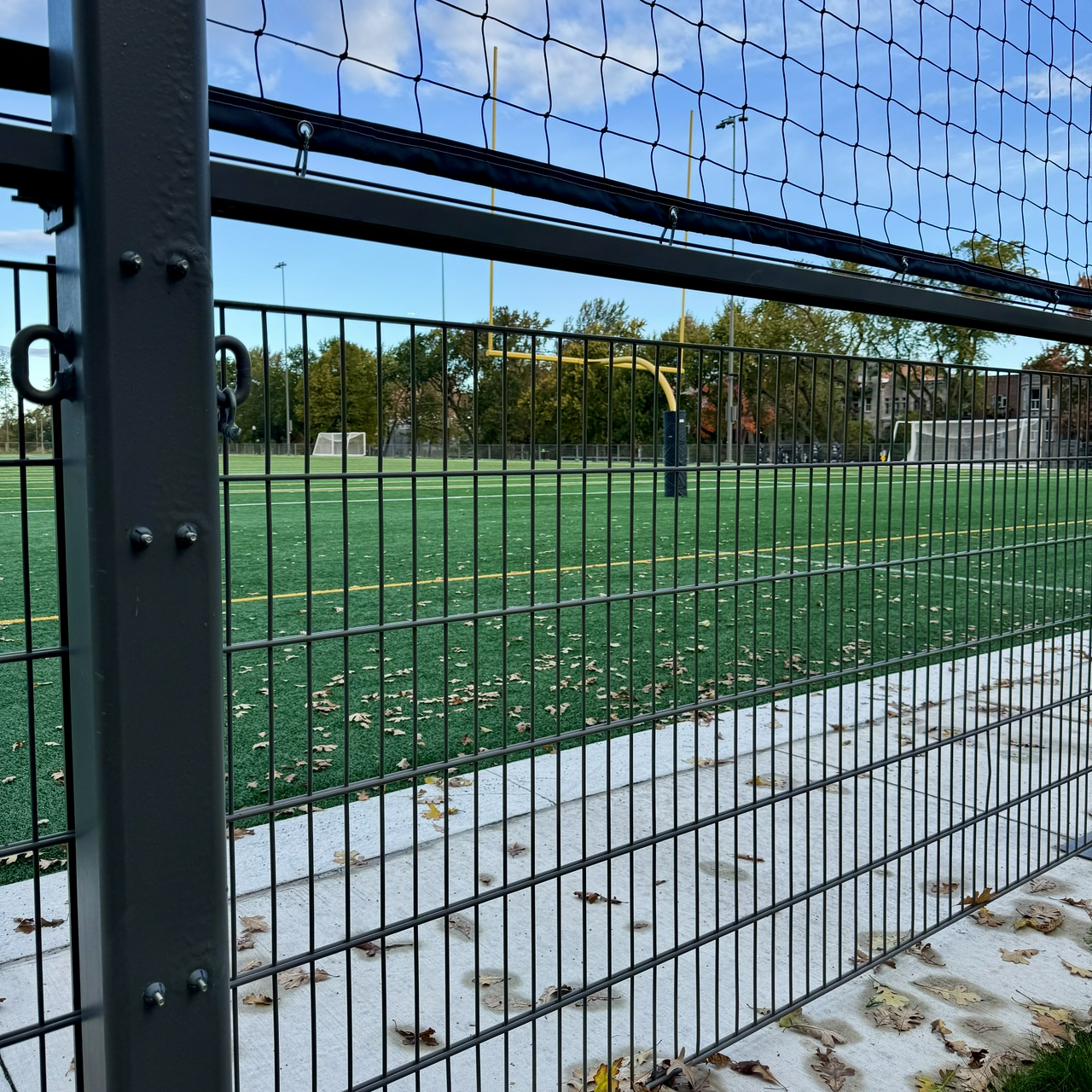 A view through a black metal fence onto a green football field. There is a yellow football goalpost in the background and fallen leaves on the ground.