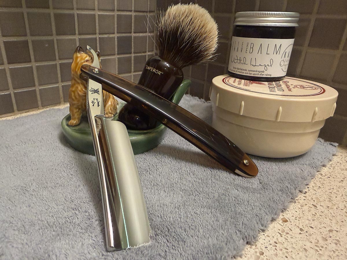 On a gray microfiber towel a straight razor with blade open about 45 degrees leans next to a bottle of aftershave and a jar of shave soap. A shave brush rests bristles-up in a green Wade ceramic pipe rest that features a brown Cairn Terrier.