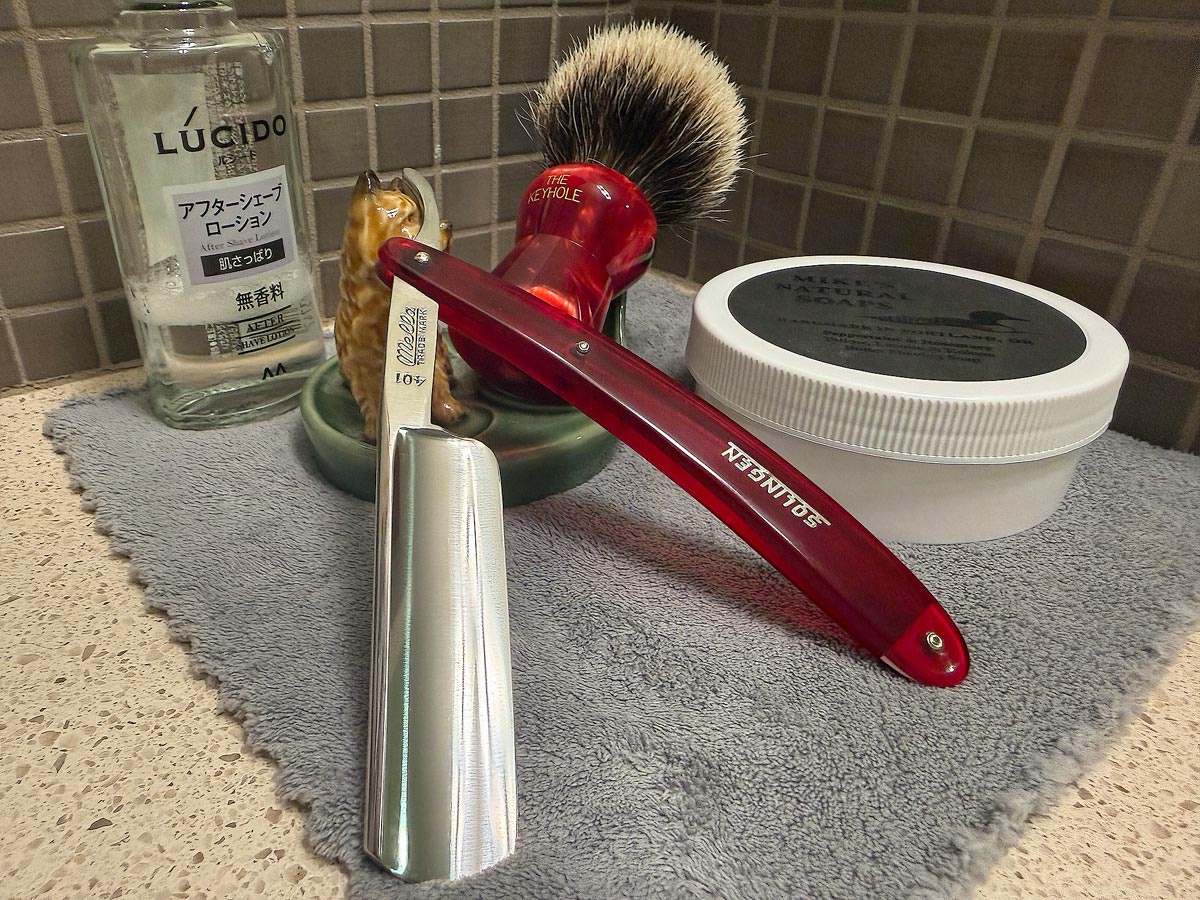 On a gray microfiber towel a straight razor with blade open about 45 degrees leans next to a bottle of aftershave and a jar of shave soap. A shave brush rests bristles-up in a green Wade ceramic pipe rest that features a brown Cairn Terrier.