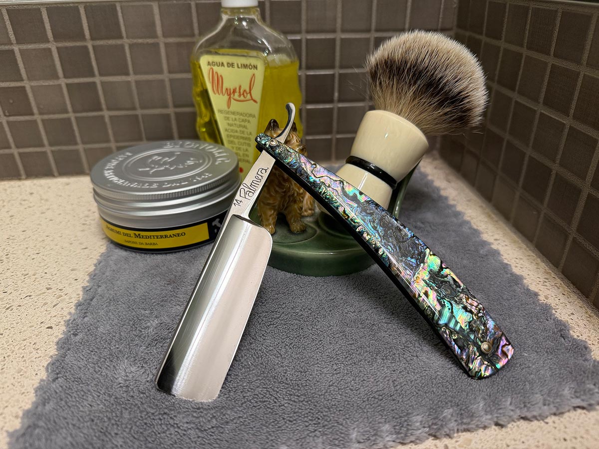 On a gray microfiber towel a straight razor with blade open about 45 degrees leans next to a bottle of aftershave and a jar of shave soap. A shave brush rests bristles-up in a green Wade ceramic pipe rest that features a brown Cairn Terrier.
