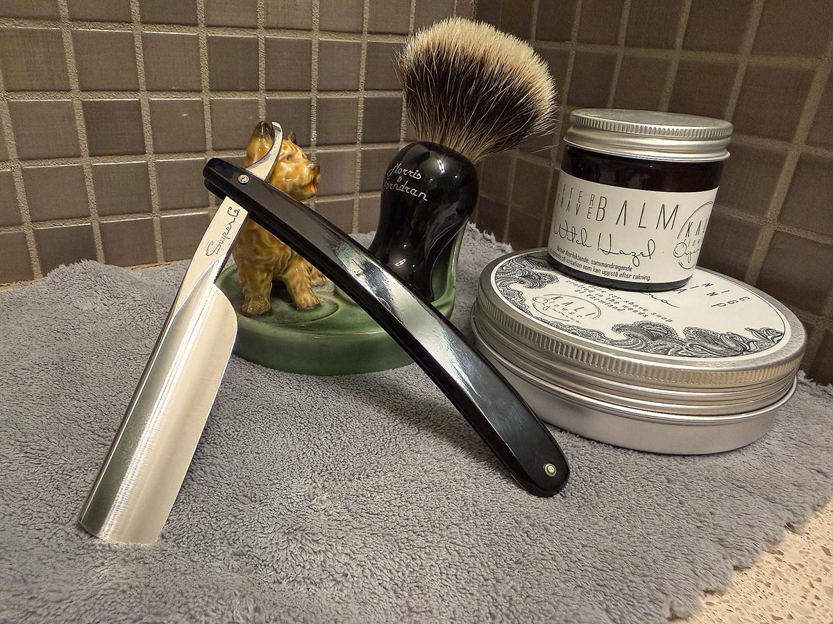 On a gray microfiber towel a straight razor with blade open about 45 degrees leans next to a bottle of aftershave and a jar of shave soap. A shave brush rests bristles-up in a green Wade ceramic pipe rest that features a brown Cairn Terrier.
