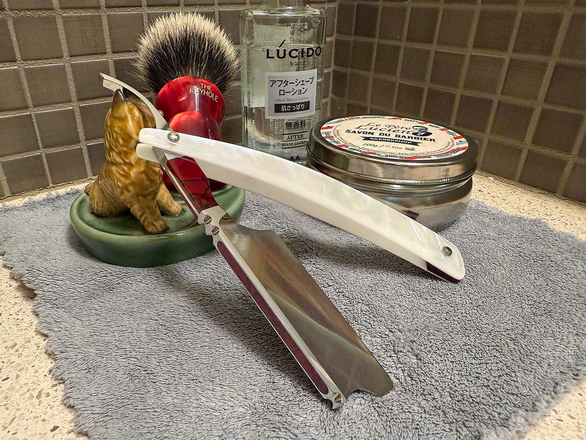 On a gray microfiber towel a straight razor with blade open about 45 degrees leans next to a bottle of aftershave and a jar of shave soap. A shave brush rests bristles-up in a green Wade ceramic pipe rest that features a brown Cairn Terrier.