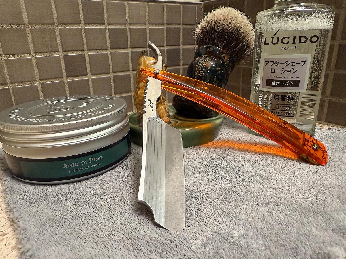 On a gray microfiber towel a straight razor with blade open about 45 degrees leans next to a bottle of aftershave and a jar of shave soap. A shave brush rests bristles-up in a green Wade ceramic pipe rest that features a brown Cairn Terrier.