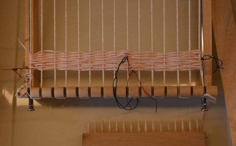 un telar en el que hay un tejido rosa palo con hilos rojo, azul y blanco anudados, mostrando cuentas doradas y azules