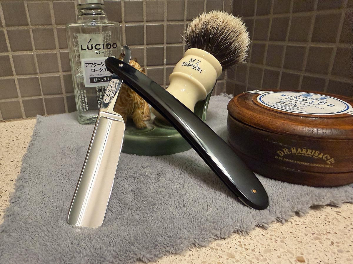 On a gray microfiber towel a straight razor with blade open about 45 degrees leans next to a bottle of aftershave and a jar of shave soap. A shave brush rests bristles-up in a green Wade ceramic pipe rest that features a brown Cairn Terrier.