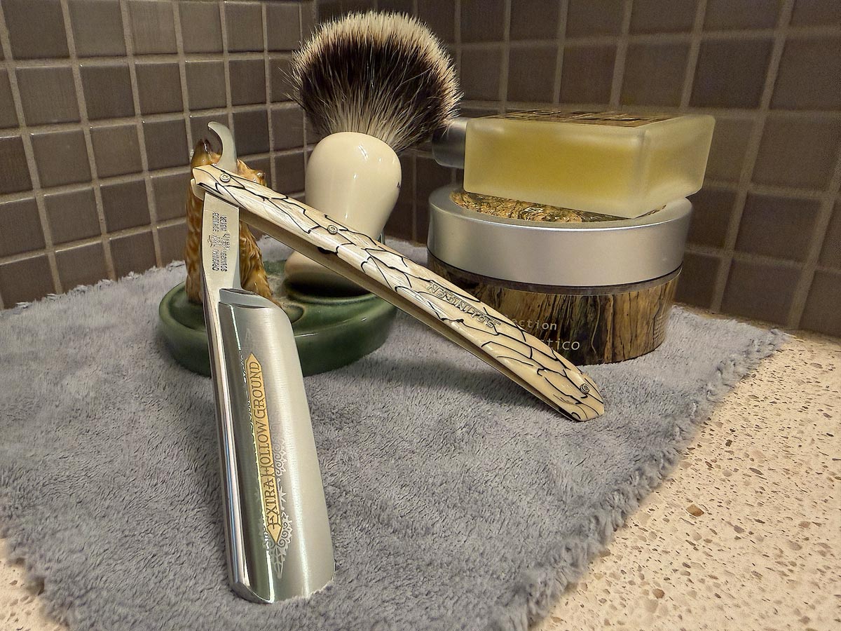 On a gray microfiber towel a straight razor with blade open about 45 degrees leans next to a bottle of aftershave and a jar of shave soap. A shave brush rests bristles-up in a green Wade ceramic pipe rest that features a brown Cairn Terrier.