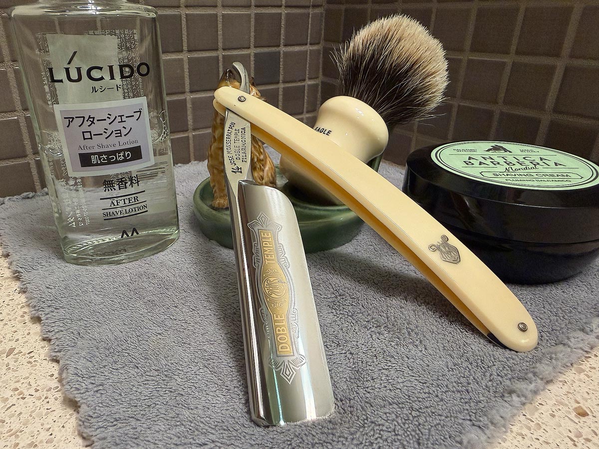 On a gray microfiber towel a straight razor with blade open about 45 degrees leans next to a bottle of aftershave and a jar of shave soap. A shave brush rests bristles-up in a green Wade ceramic pipe rest that features a brown Cairn Terrier.