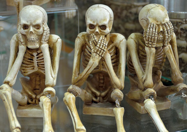 Three skeleton statues sitting on a clear surface. The left skeleton has its hands on its ears. The middle skeleton has its hands covering its mouth. The third skeleton has its hands on its eyes.