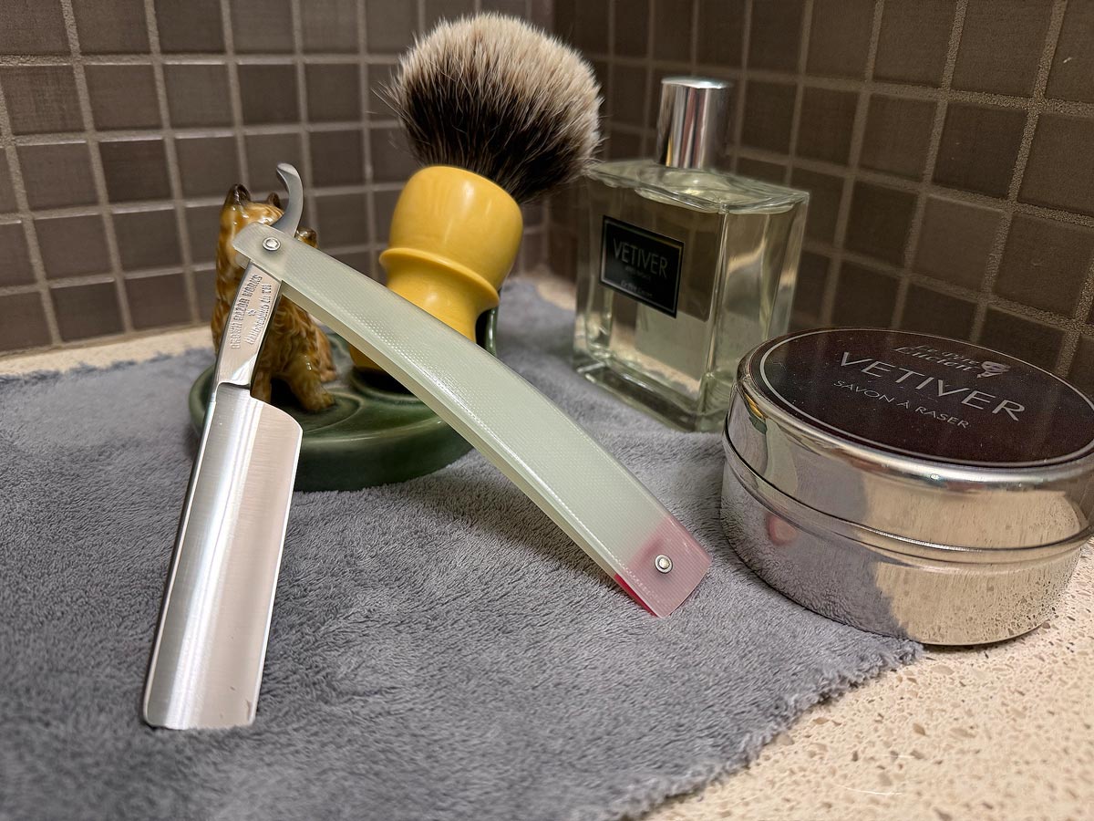 On a gray microfiber towel a straight razor with blade open about 45 degrees leans next to a bottle of aftershave and a jar of shave soap. A shave brush rests bristles-up in a green Wade ceramic pipe rest that features a brown Cairn Terrier.